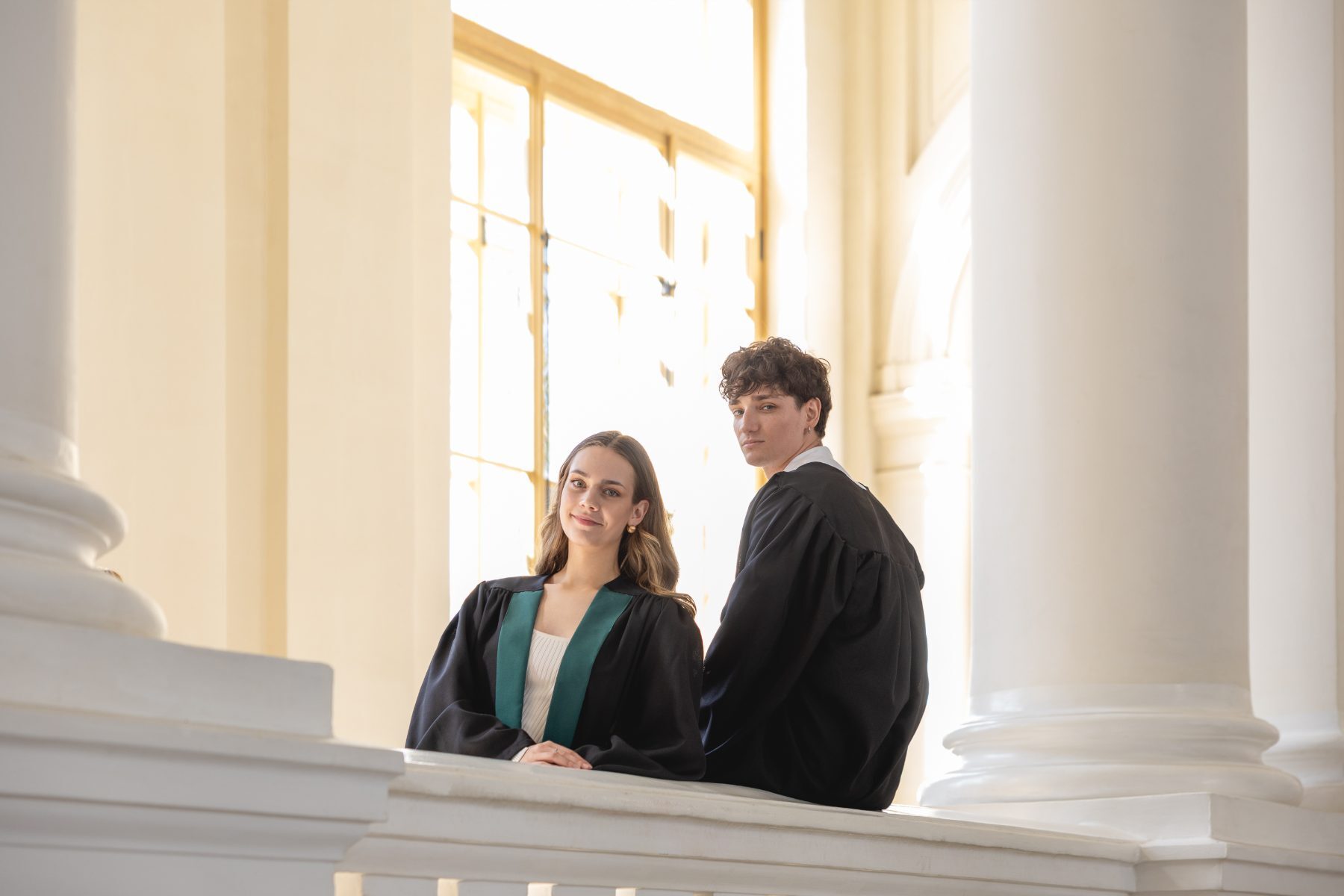 Two graduates in black robes seated on a balustrade between white columns