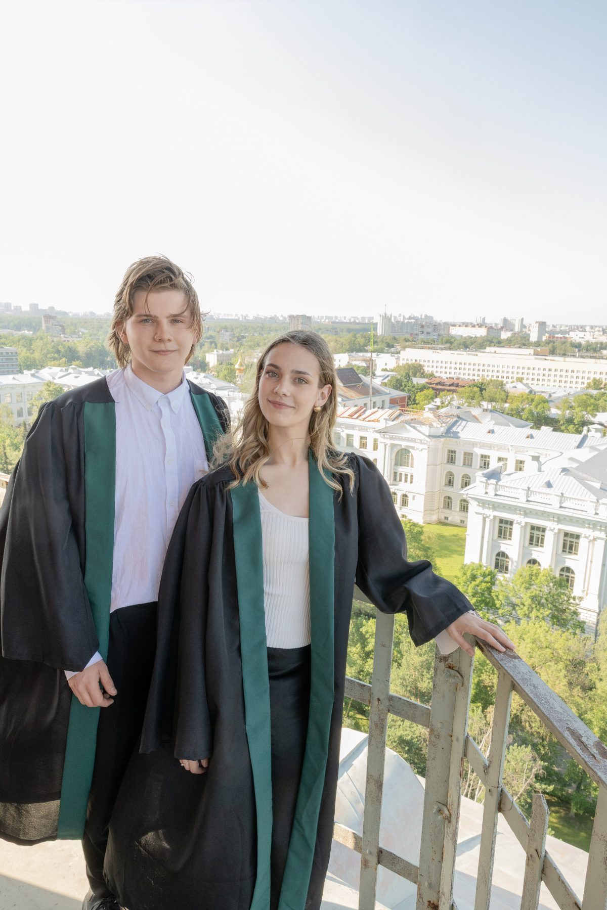 Two graduates in green-trimmed robes on a rooftop above the city