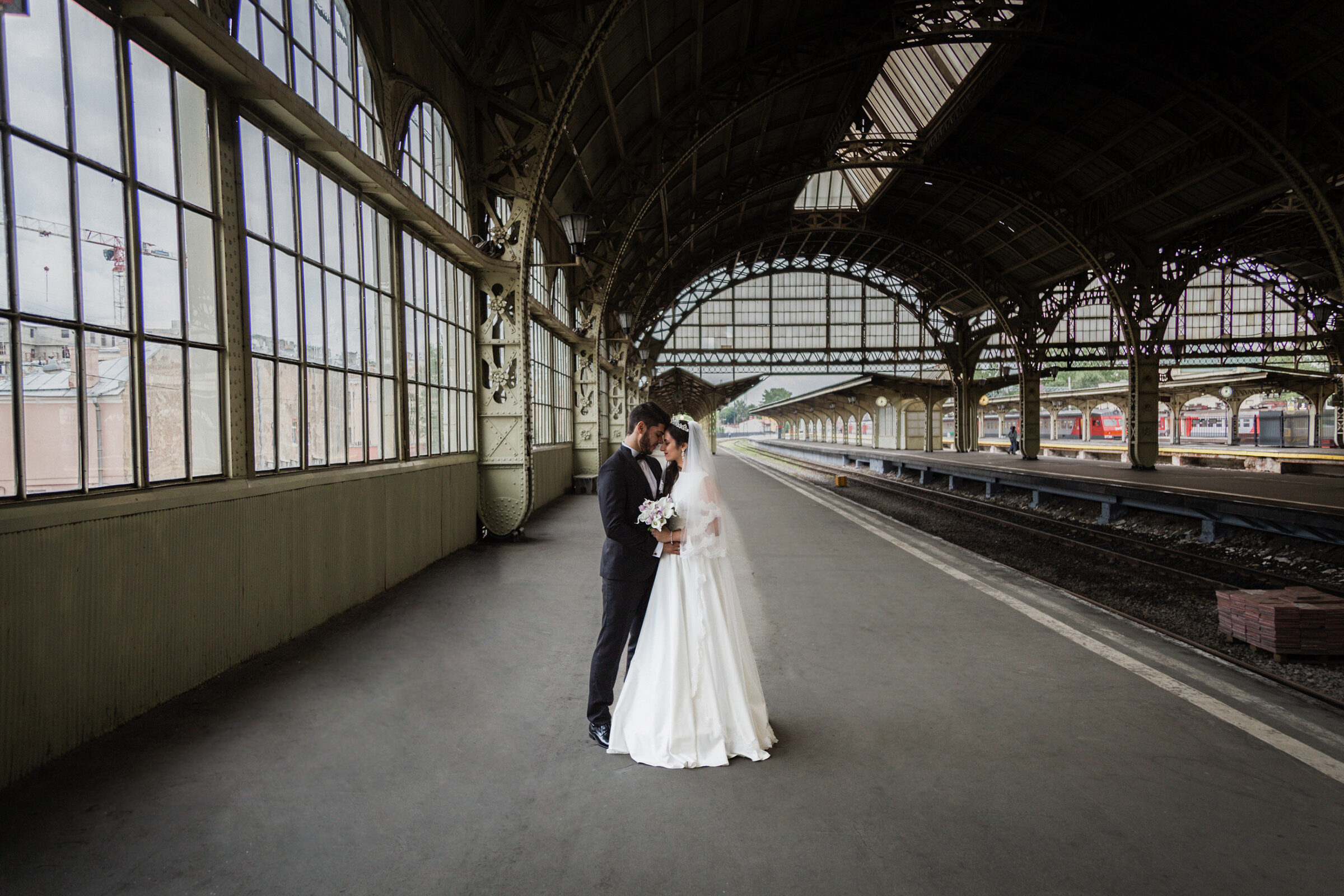 Bride and groom forehead to forehead under the iron arches of a railway station platform