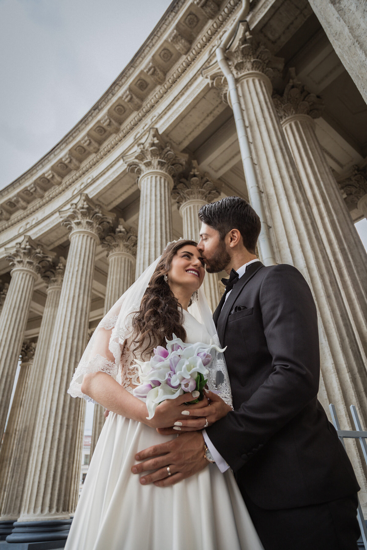 Groom kissing the bride’s forehead beneath a curved stone colonnade