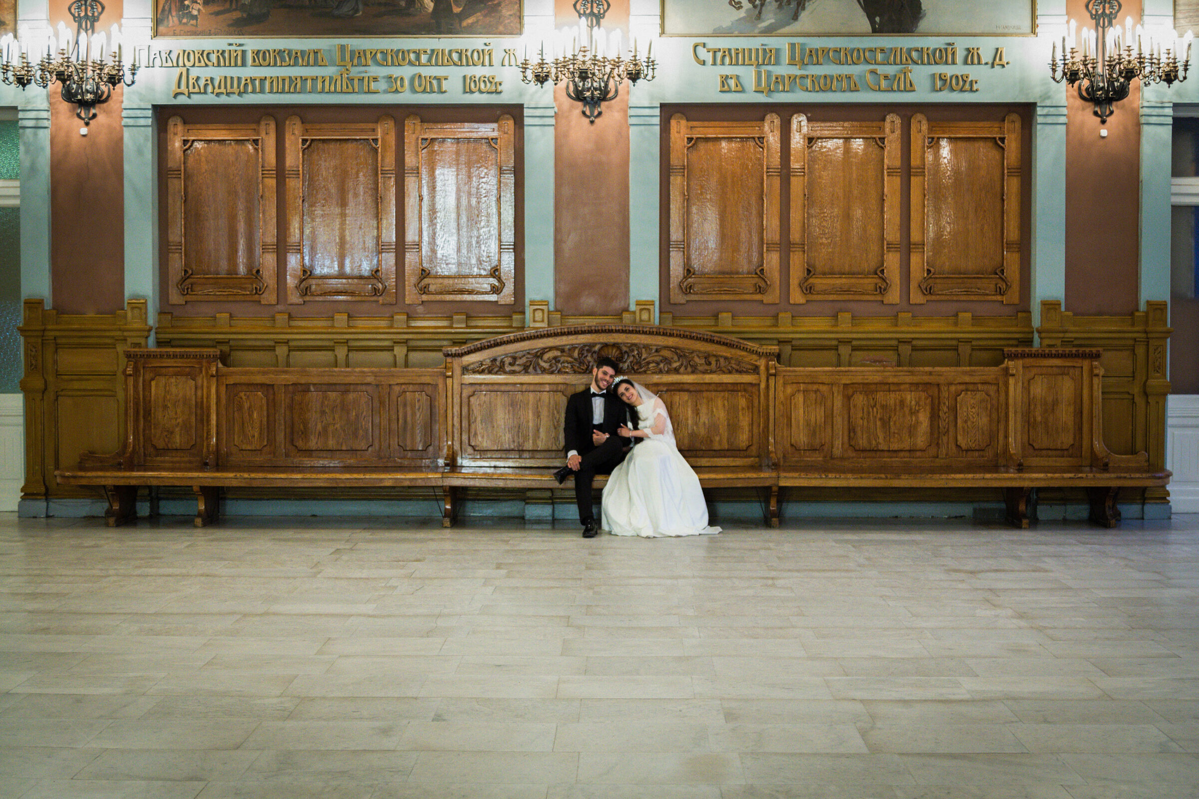 Couple sitting close on a long carved wooden bench in a historic station hall