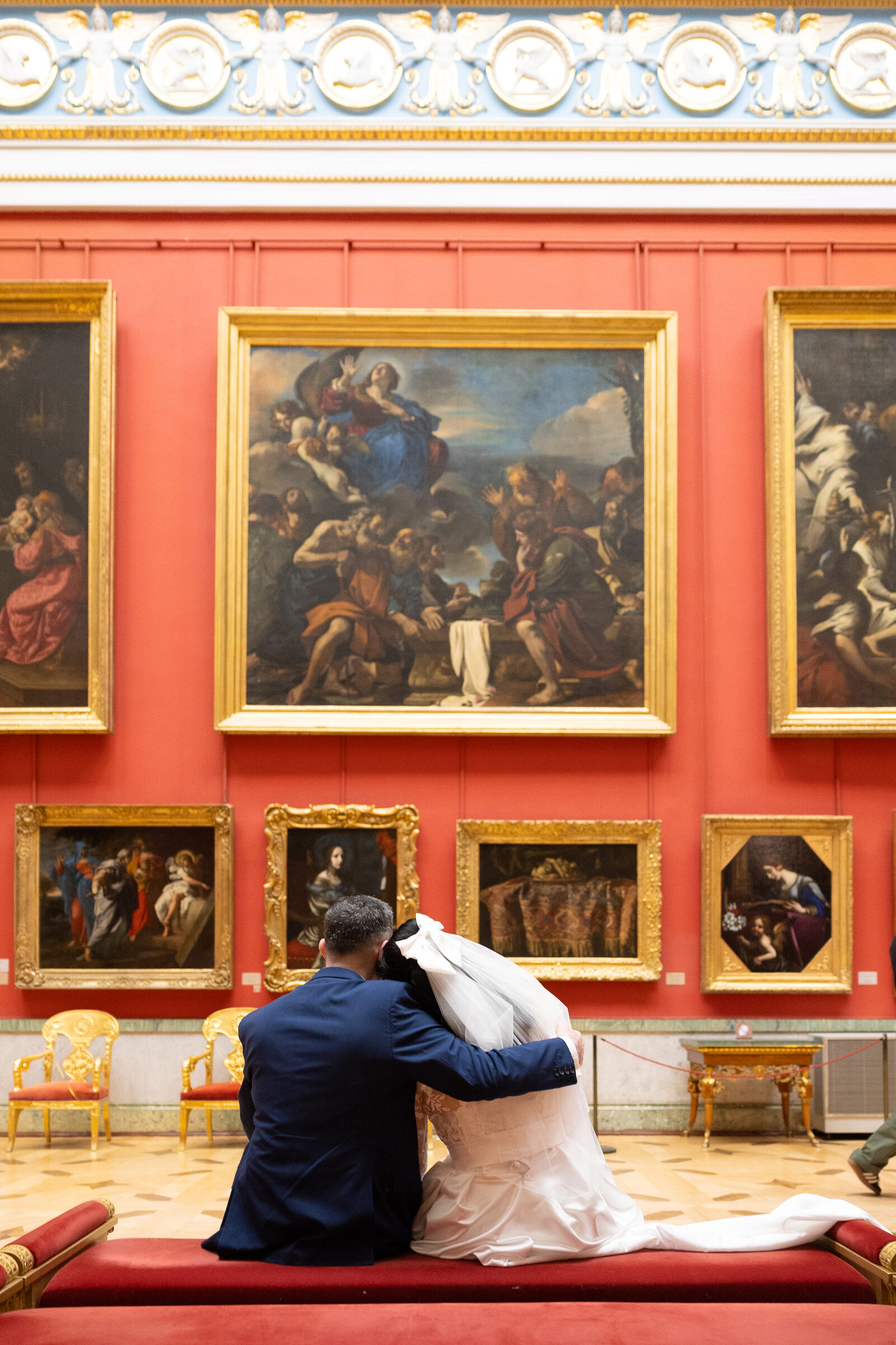 Couple seen from behind on a red bench, her head on his shoulder, facing old paintings