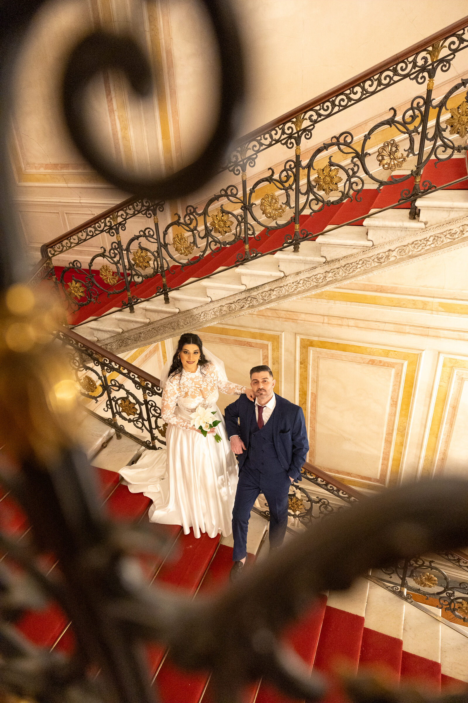 Couple on a red-carpeted staircase, framed by a blurred wrought-iron scroll