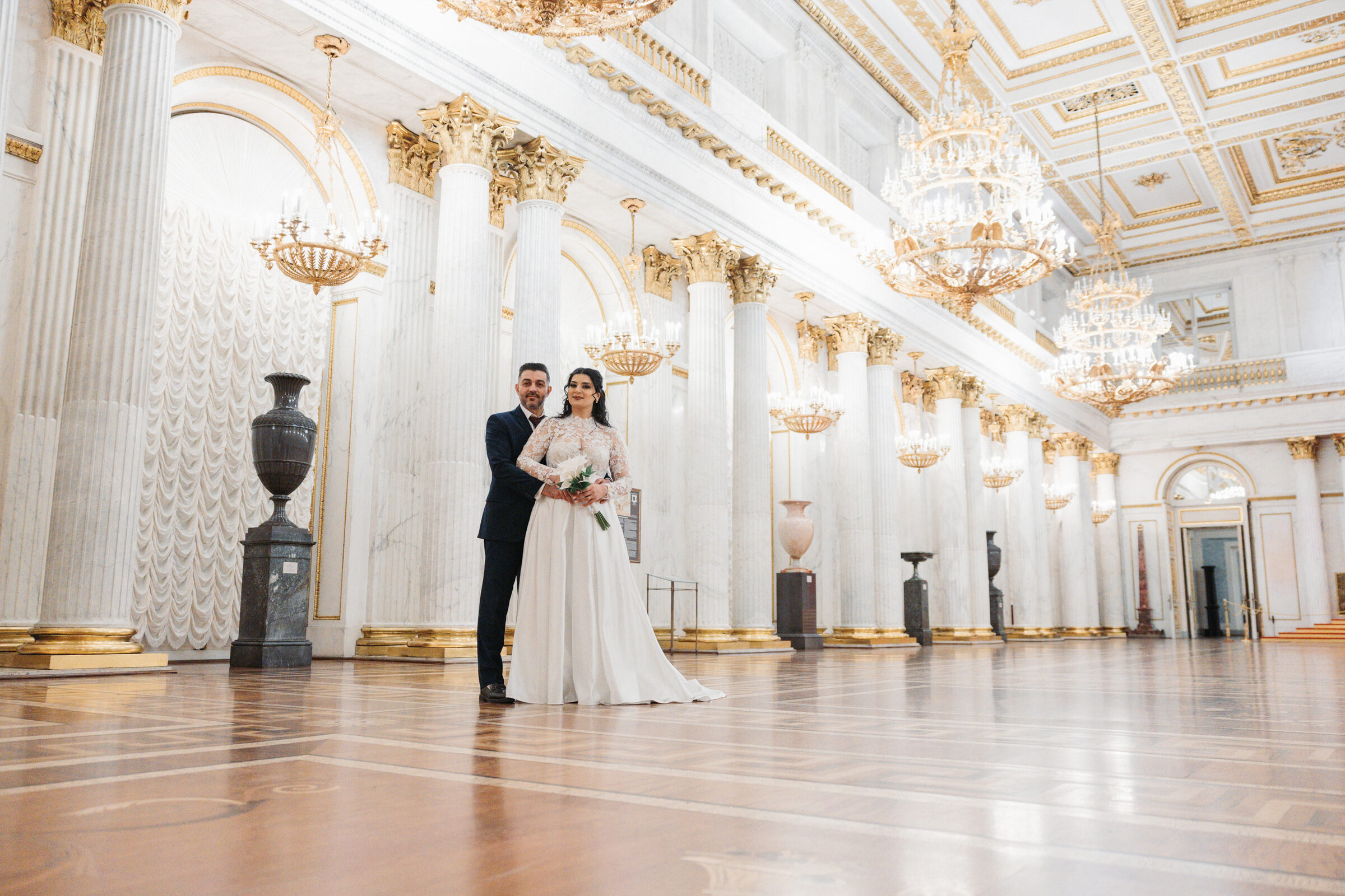 Couple standing together in a white marble hall under gold chandeliers