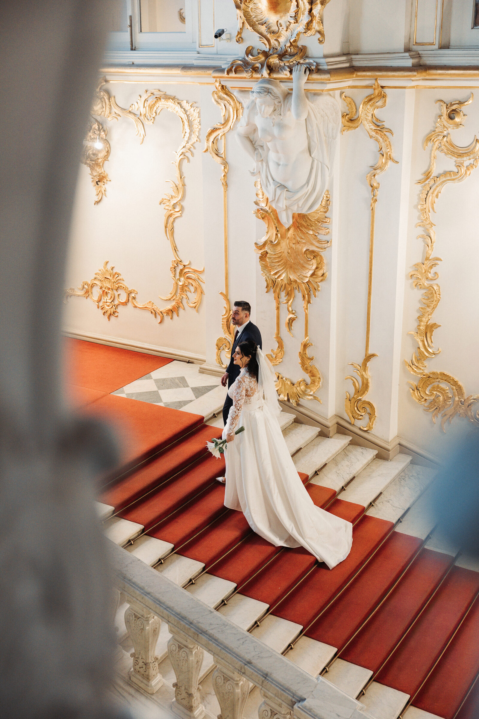 Overhead view of the couple descending a red-carpeted staircase past gold ornament