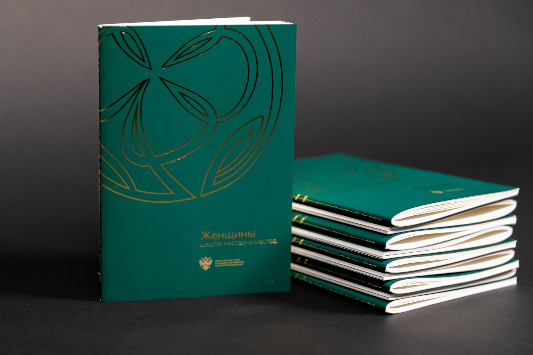 Green booklets with gold foil covers standing and stacked against a dark background