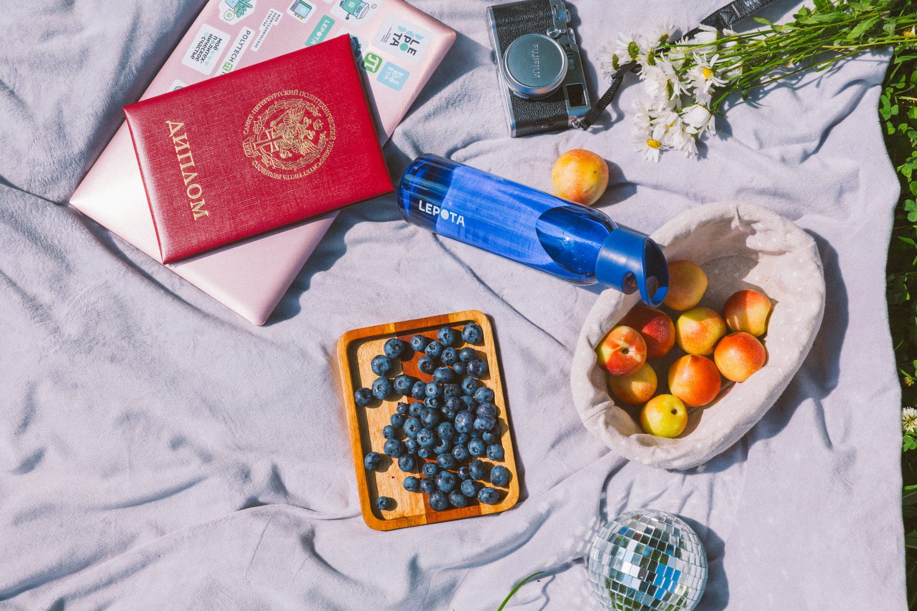 Flatlay on a picnic blanket: a branded blue bottle, red diploma, camera, blueberries and peaches