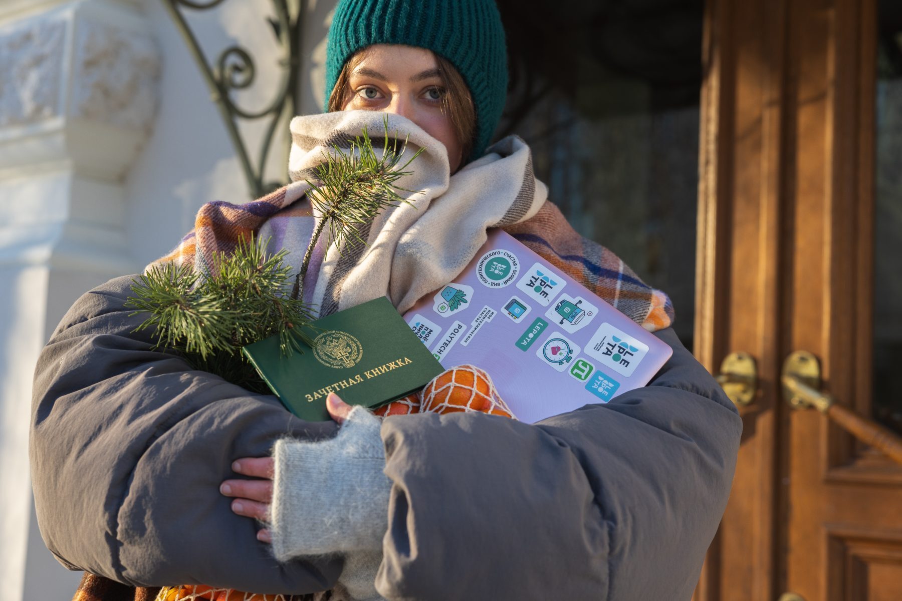 Person bundled in a scarf and hat holding a sticker-covered laptop, record book and mandarins outdoors in winter