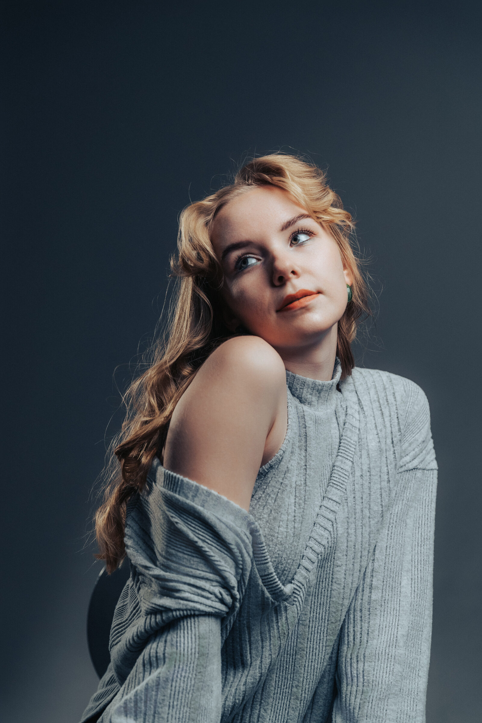 Woman in a grey cut-out sweater looking up over her shoulder against a dark slate backdrop