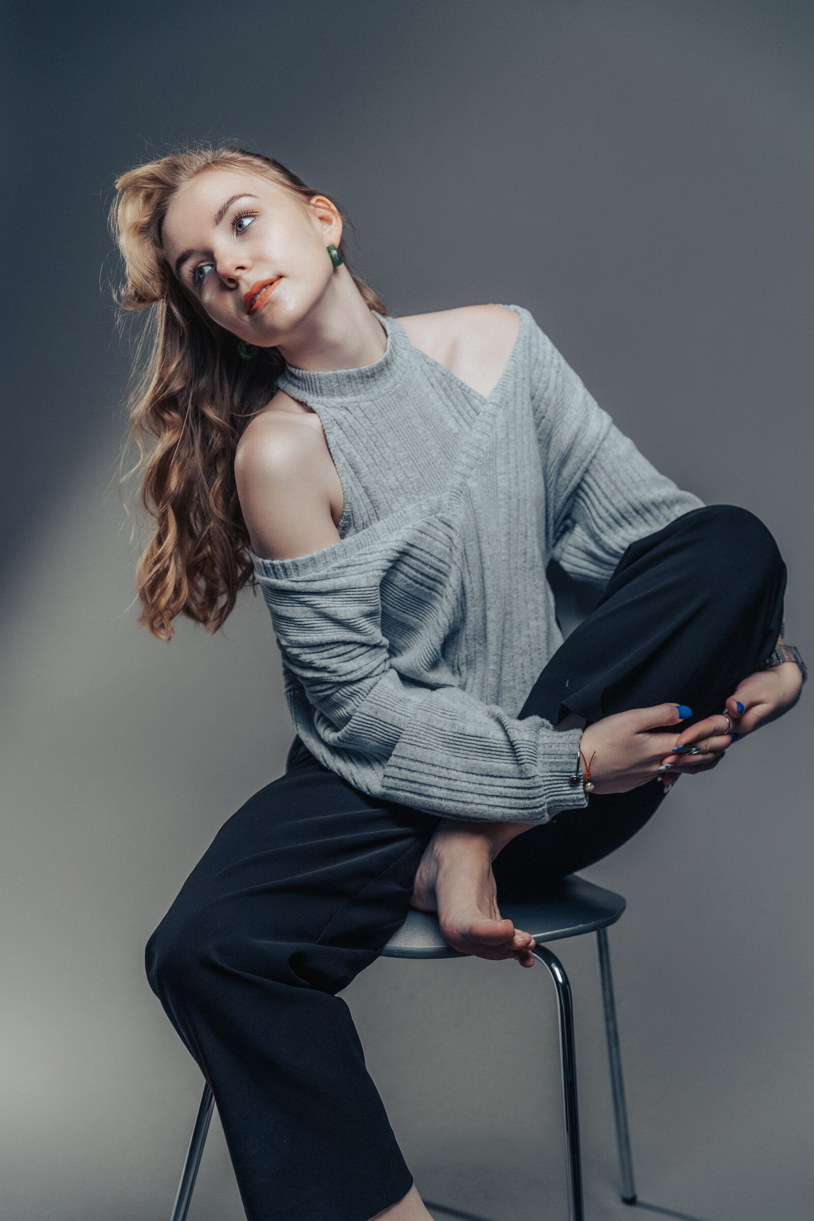 Full-length studio portrait of a woman perched barefoot on a metal stool, hugging her knee